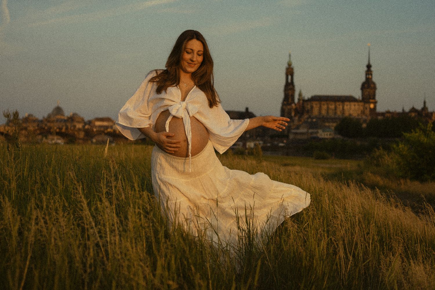 Portrait Fotoshooting Elbwiesen Dresden - Schwangere Frau zeigt nackten Bauch und schwingt ihren weißen Rock bei Sonnenuntergang