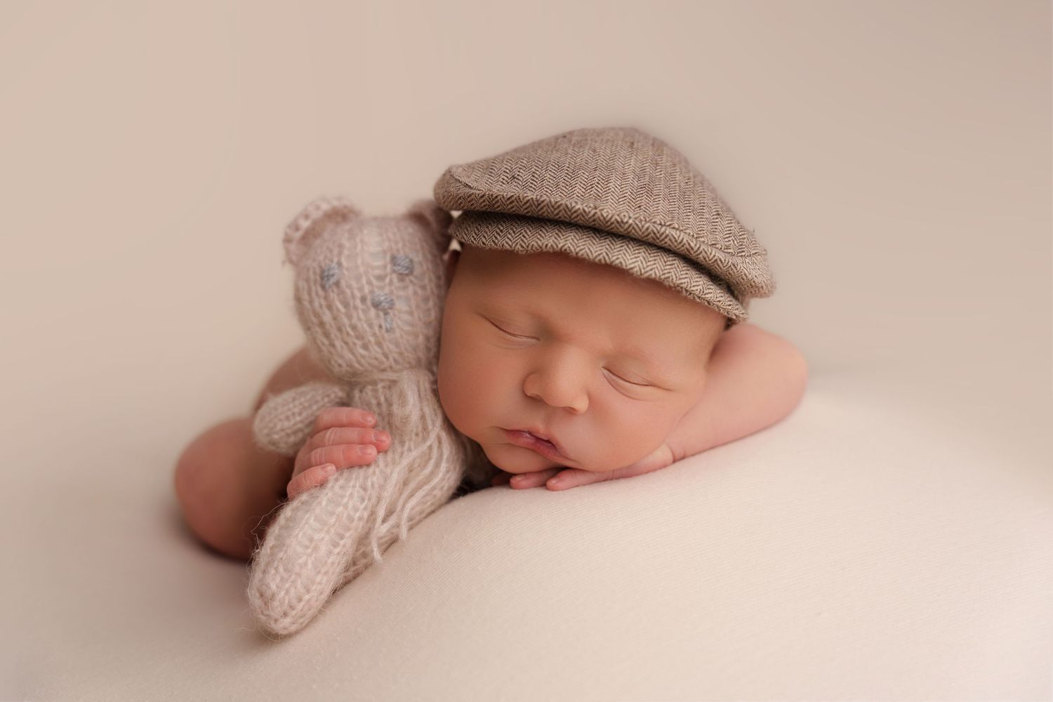 Sleeping newborn wearing knit cap cuddling with teddy bear on neutral backdrop.