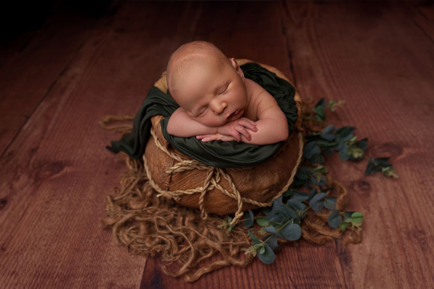 Sleeping newborn nestled in rustic wooden bowl with green vine decorations.