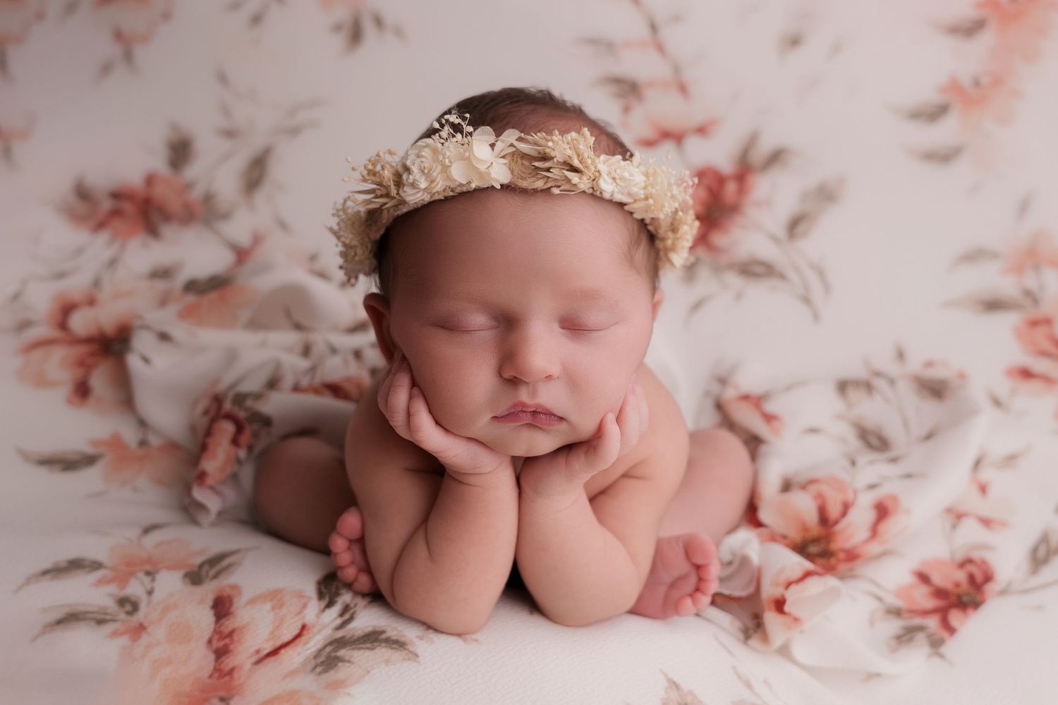 Sleeping newborn poses on floral fabric wearing a delicate flower crown in soft lighting.