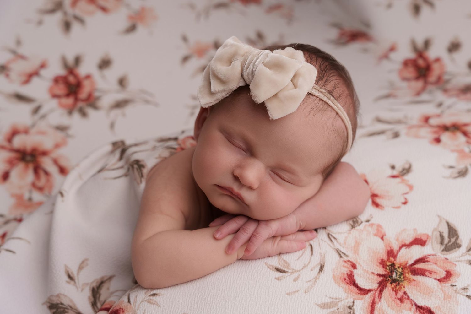 Sleeping newborn on floral bedding wearing white bow headband in soft natural light.