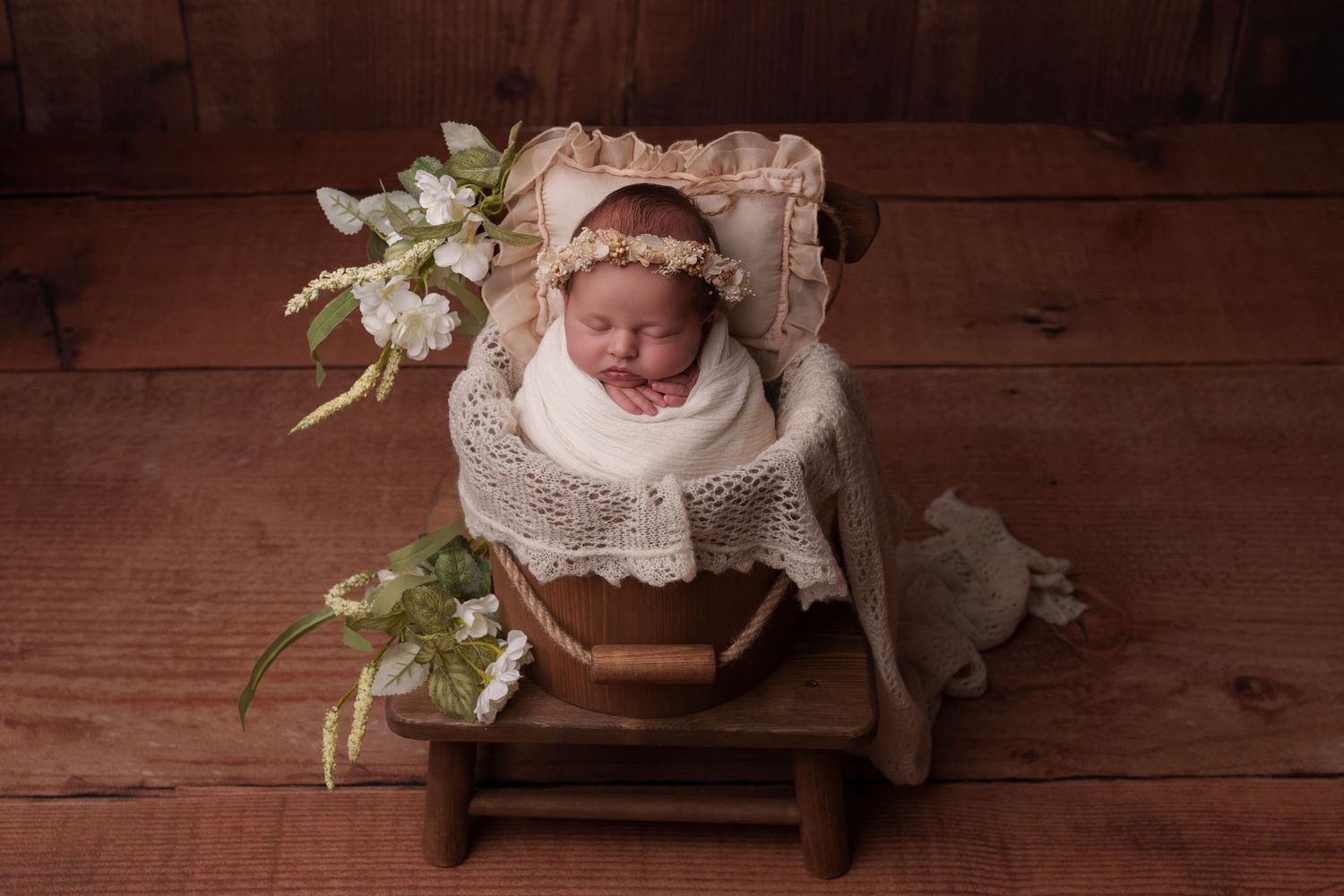 Newborn sleeping on vintage wooden chair with lace swaddle and white flowers.