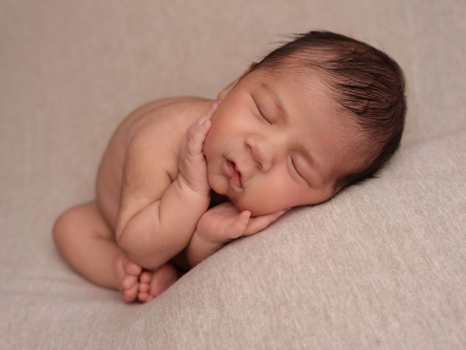Peaceful sleeping newborn baby curled up on soft beige blanket in artistic portrait pose.