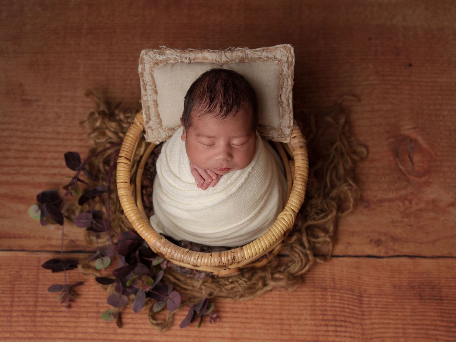 Sleeping newborn in white wrap posed in basket with purple flowers.