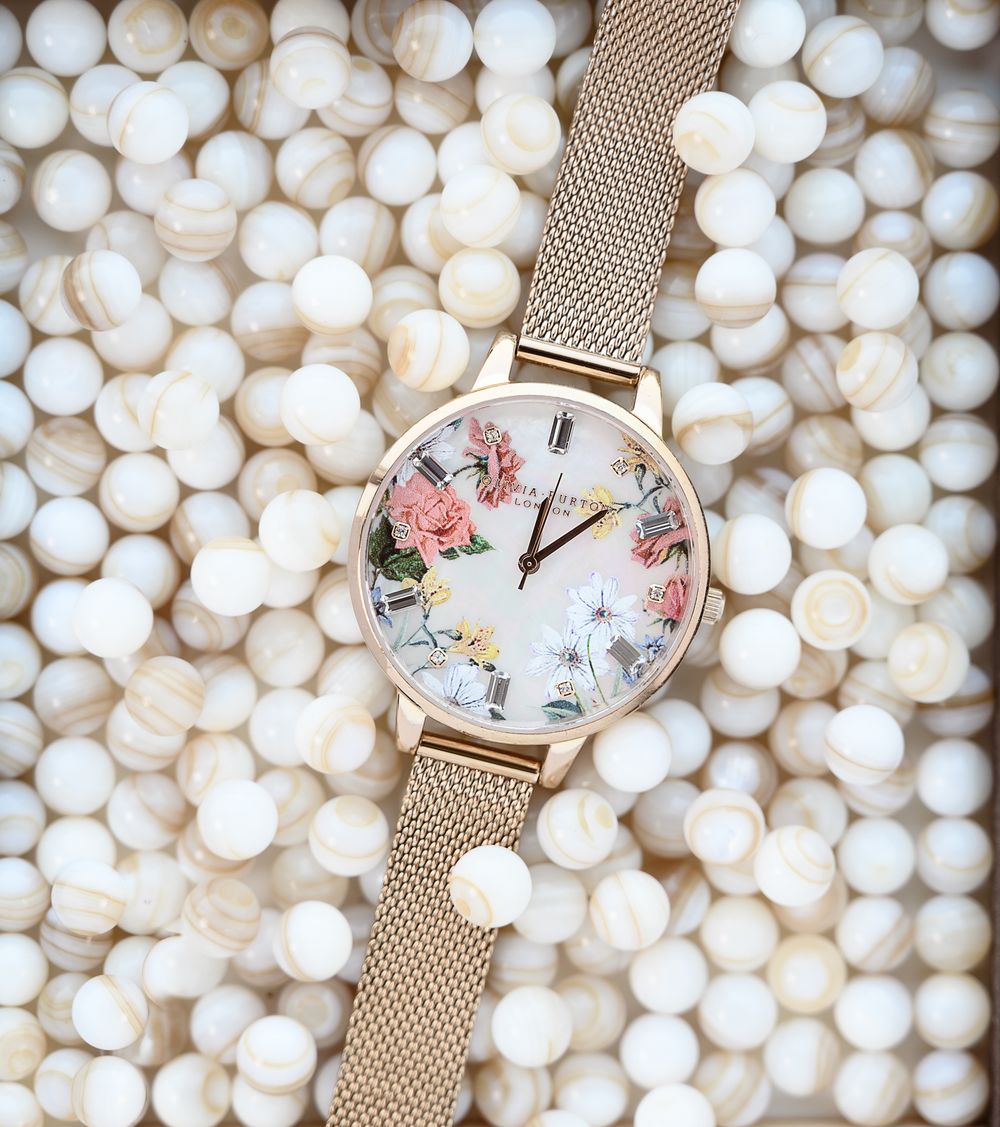 Gros plan d'une montre de marque au cadran doré orné de fleurs sur un lit de perles, couleurs pastels, ambiance chic et raffinée.