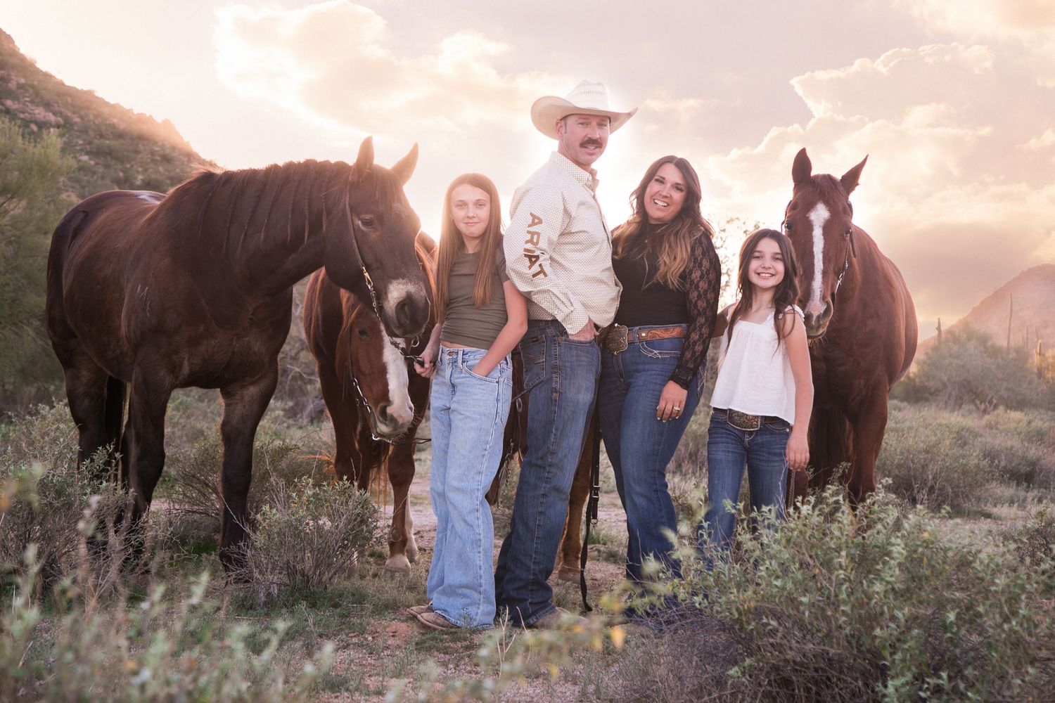 Family of four posing with two brown horses in a desert landscape at golden hour.