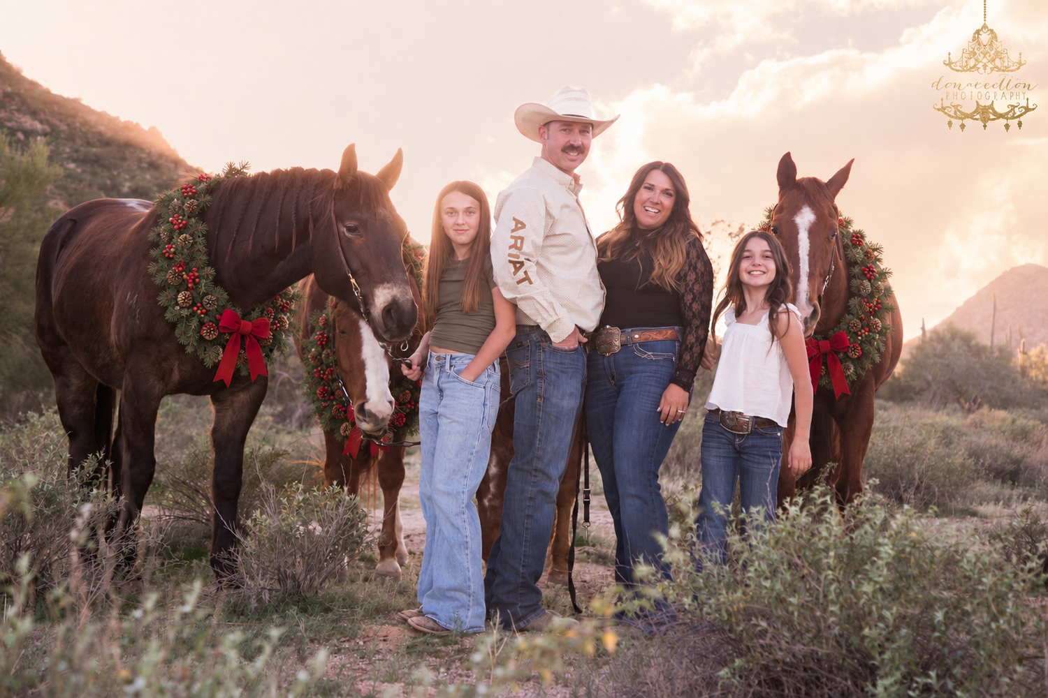 Family of four posing with two brown horses in a desert landscape at golden hour.