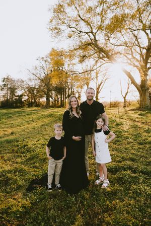 Family of four posing in golden field at sunset, mother in black maternity gown.