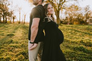 Pregnant woman in black maternity gown leans back against partner at golden hour outdoors.