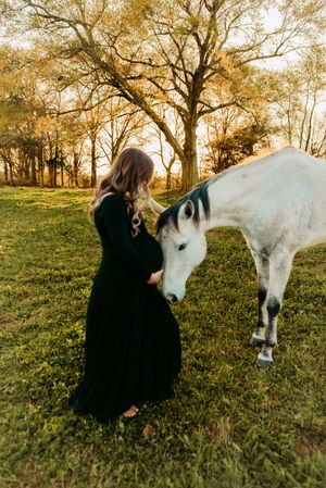 Pregnant woman in black gown gently touching nose of white horse in autumn field.