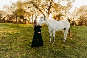 Smiling pregnant woman in black gown standing beside white horse at golden hour outdoors.