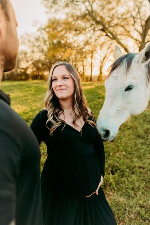 Glowing pregnant woman in black dress smiling beside white horse at warm sunset outdoors.