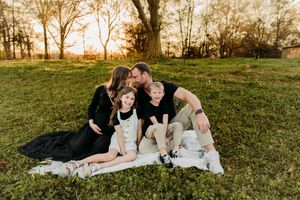 Family of four sitting on blanket at golden hour sunset in park, parents kissing while children smile.