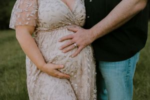 Close-up of couple's hands cradling pregnant belly in floral lace maternity dress outdoors.