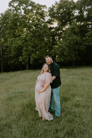 Smiling pregnant couple embracing in green meadow, woman in floral lace gown at golden hour.