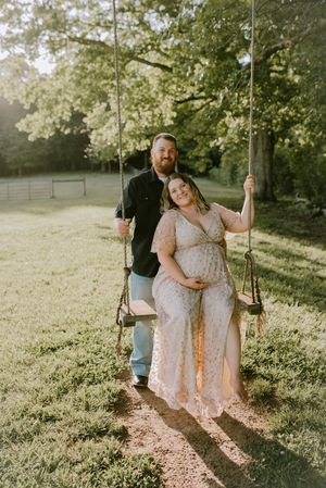 Pregnant woman in lace dress sitting on rope swing with partner standing behind in sunny backyard.