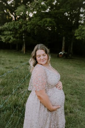 Pregnant woman in lace blush gown cradling baby bump in sunlit green field.