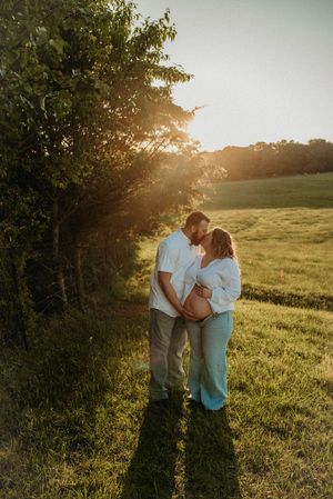 Couple sharing tender kiss at golden hour in lush green field, woman showing baby bump.