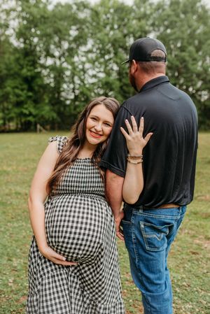 Smiling pregnant woman in gingham dress holding baby bump beside her partner outdoors in green field.