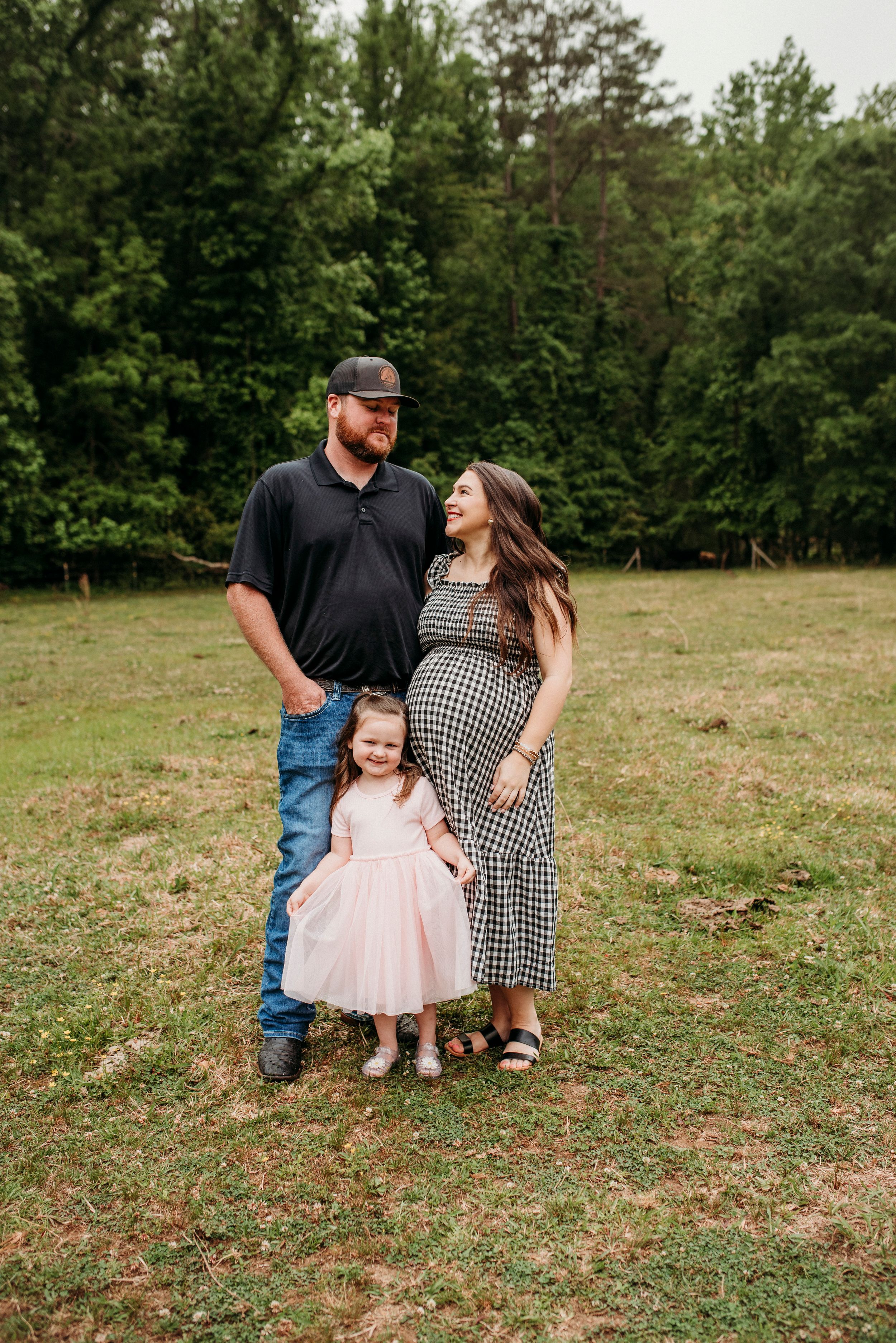 Expecting family with toddler daughter posing in grassy field, mother wearing black gingham maternity dress.