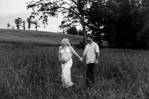 Black and white maternity photo of couple holding hands walking through tall grass field at dusk.