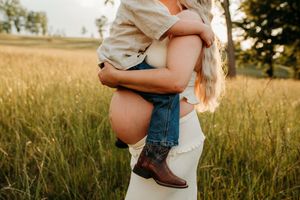 Mother holding young child wearing cowboy boots in golden grass field during warm sunset maternity session.