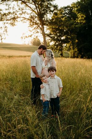 Family maternity photo in golden field, parents embracing while two young boys stand in front at sunset.