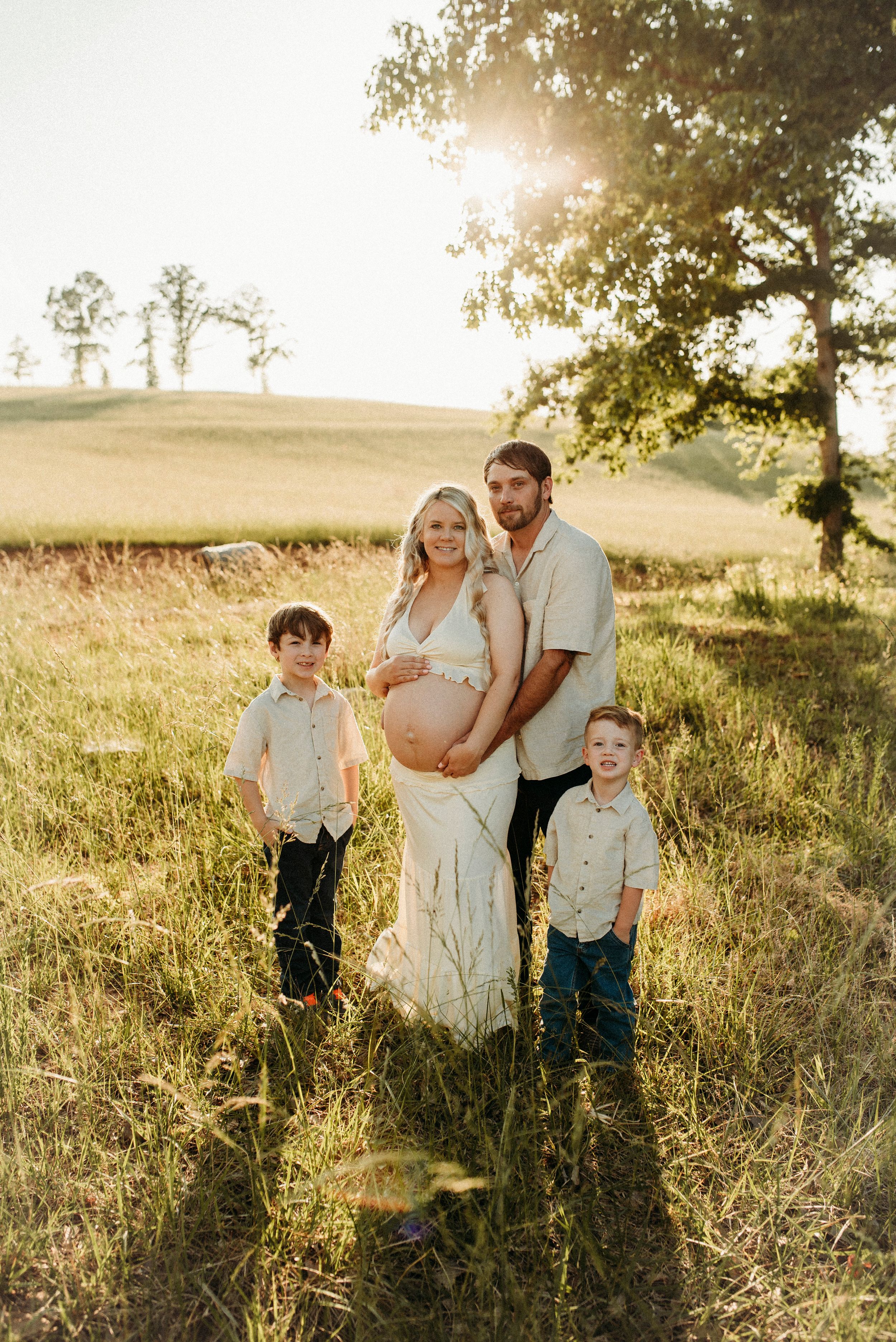 Pregnant family portrait in sunny meadow with two boys, parents holding baby bump in warm golden light.