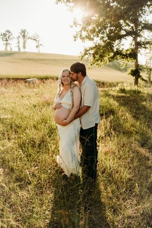 Couple sharing tender maternity moment in grassy field, man kissing pregnant woman's cheek at golden hour.