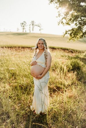 Smiling pregnant woman in white dress holding baby bump in sunlit field during golden hour outdoor shoot.