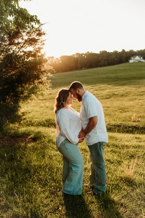 Expecting couple touching foreheads tenderly in lush green field during warm golden hour sunset session.