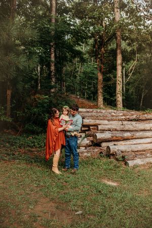 Family of three reading a book together outdoors in forest setting near stacked logs with boho styling.