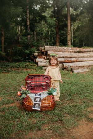 Toddler girl in yellow dress standing beside open picnic basket with ultrasound photos for pregnancy announcement.