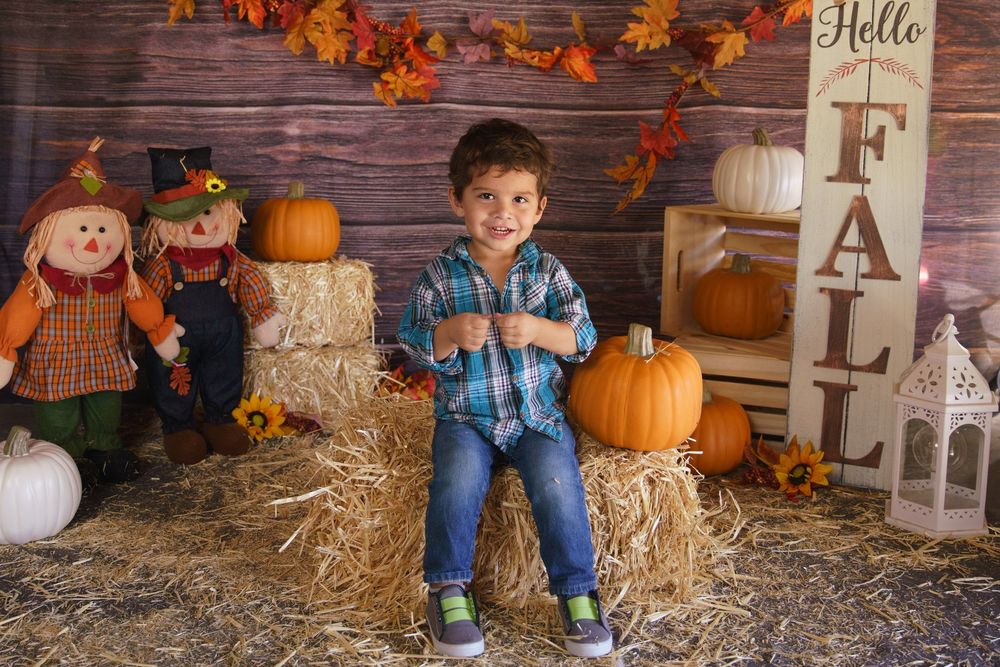 Young boy sits on hay bales surrounded by pumpkins and scarecrows in a festive fall-themed photo backdrop.