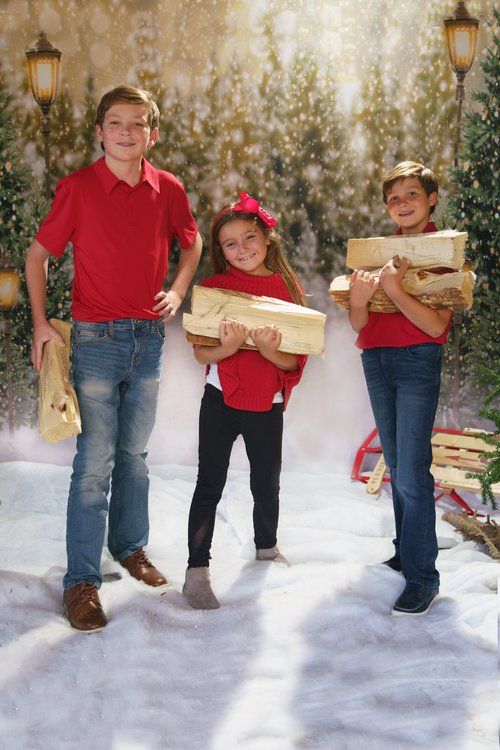 Three children in red shirts holding Christmas gifts in a snowy holiday photo backdrop with trees and lamp posts.