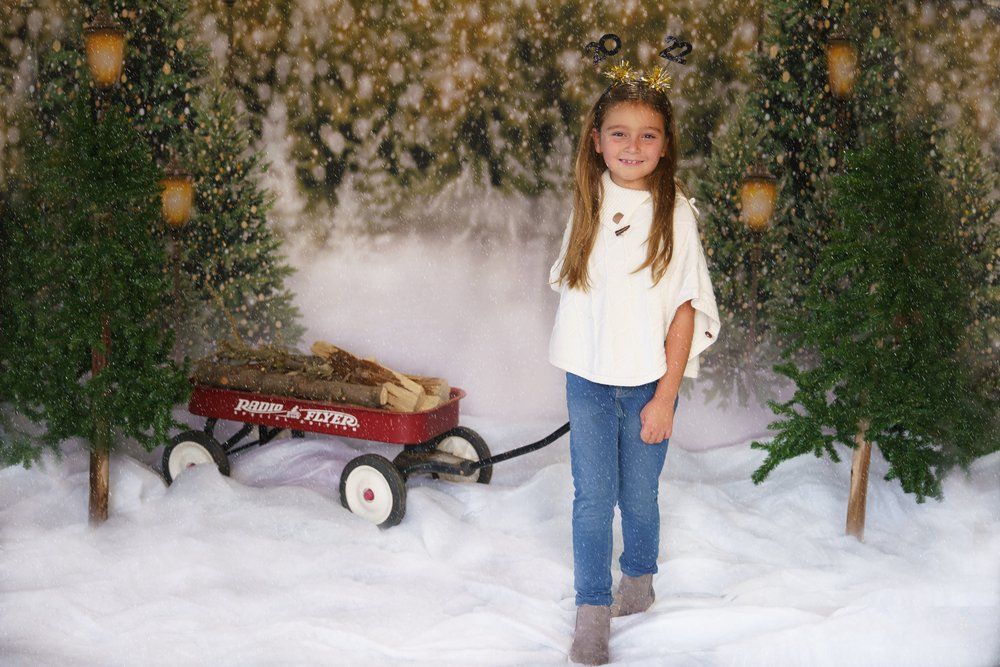 Young girl posing in winter scene with red Radio Flyer wagon, snow, and Christmas trees backdrop.