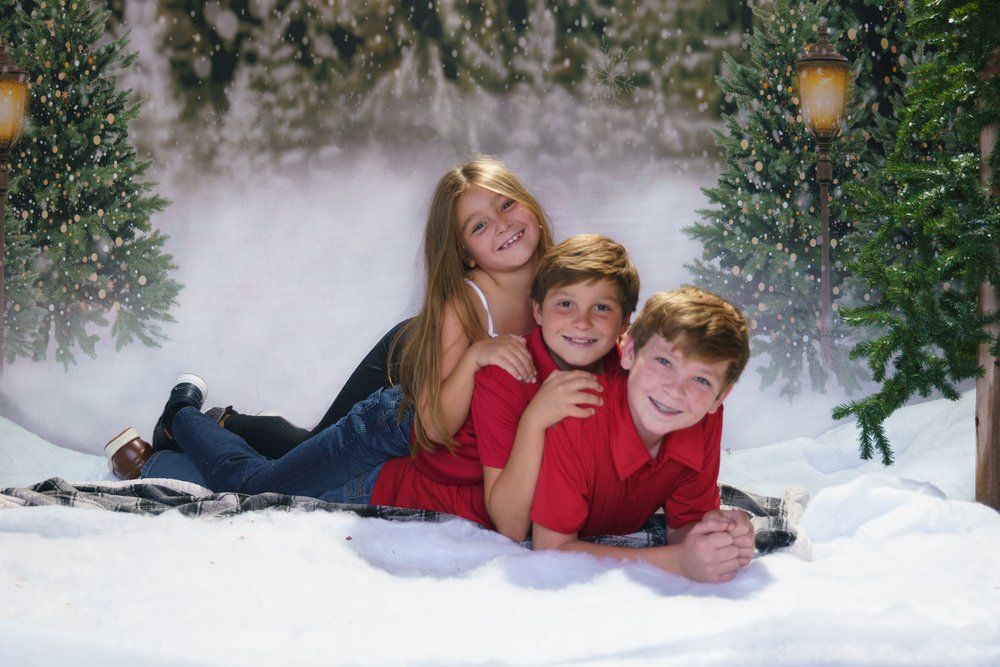 Three smiling children pose together lying in snow with a festive Christmas tree backdrop.