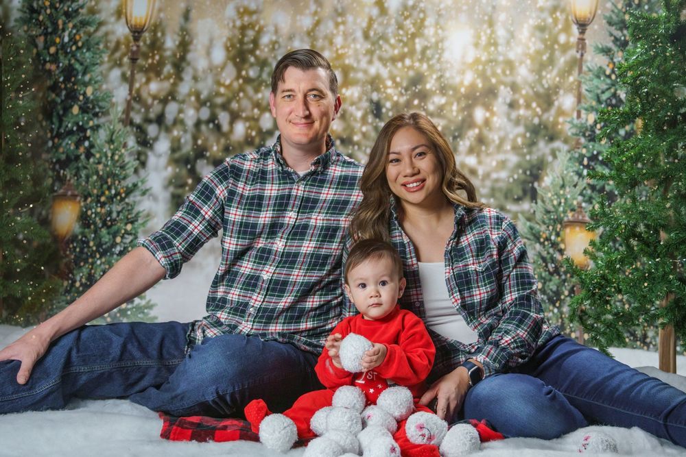 A family of three in matching plaid shirts poses with their baby in a festive Christmas photo setting.