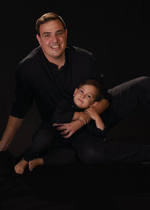 Portrait of a father and young child in matching black outfits pose together smiling against dark studio background.