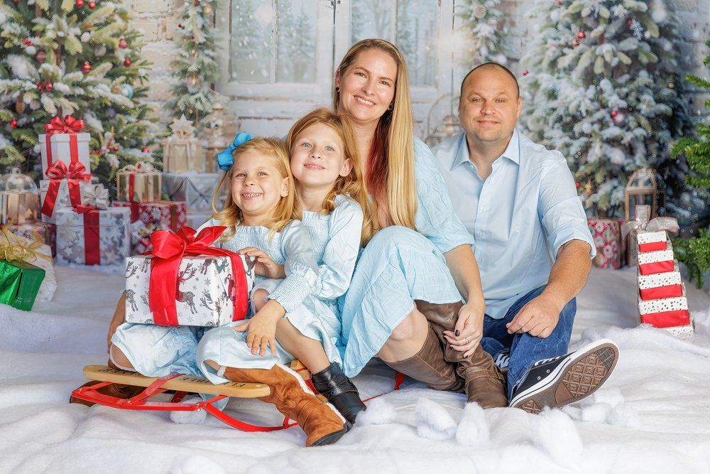 Happy family photo of four posing together in a festive Christmas photo studio with decorated trees and gifts.