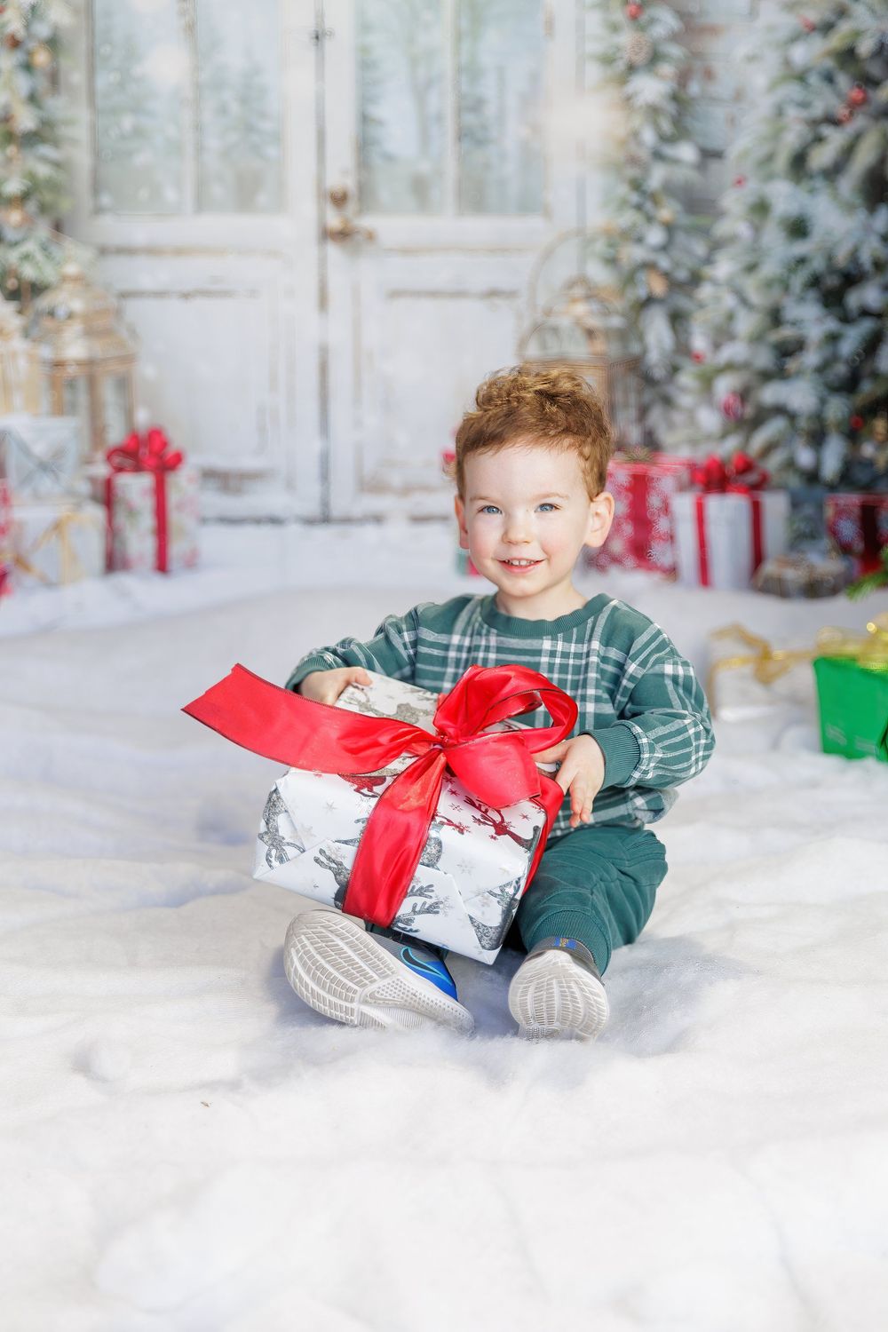 Smiling toddler boy holding a large Christmas gift with red bow, sitting on snow in festive holiday studio setting.