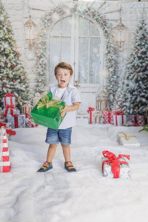 Excited young boy holding a large green Christmas gift box surrounded by presents and decorated trees.