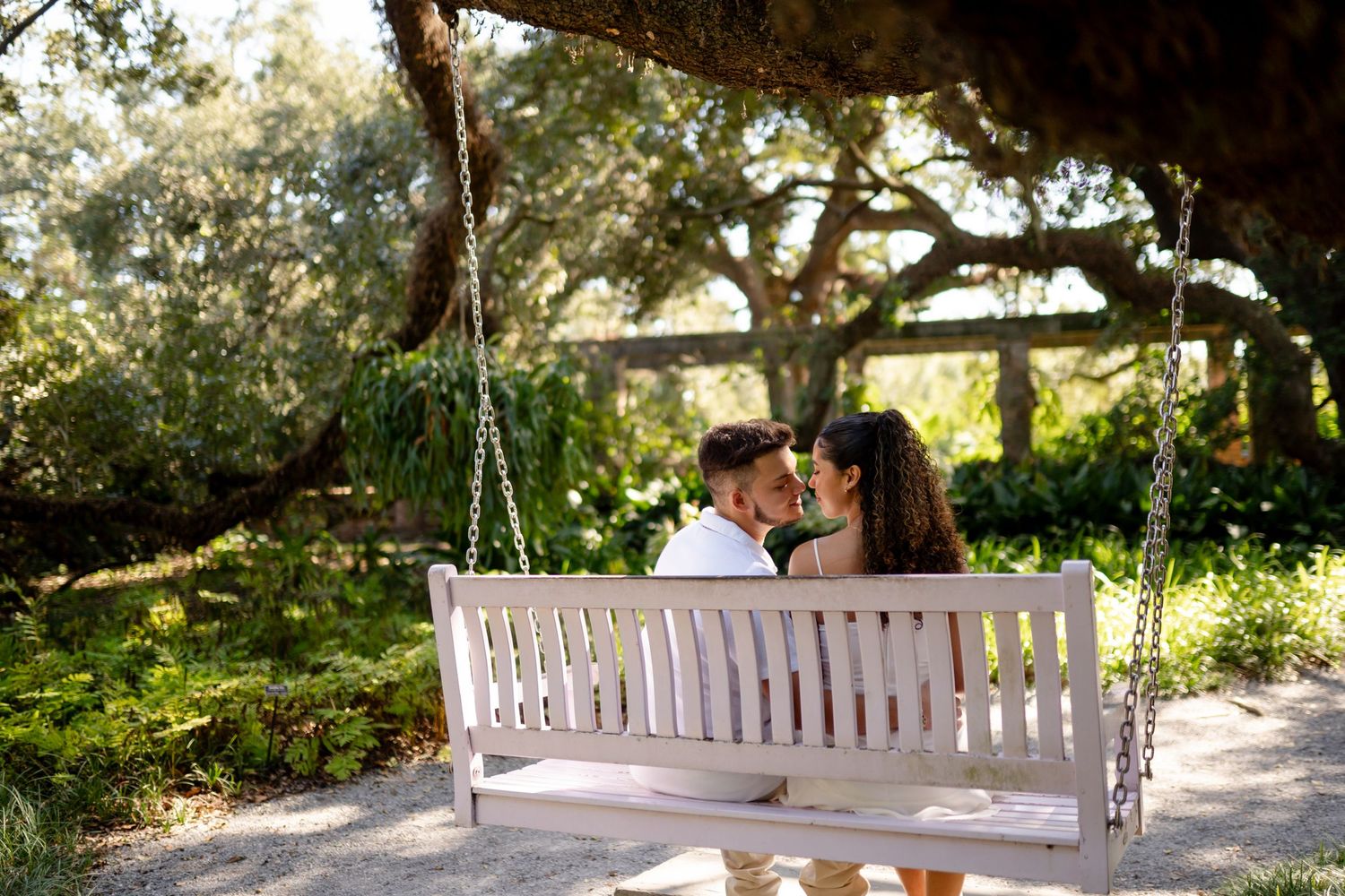 A couple shares a romantic moment on a white wooden swing beneath a shady tree in a lush garden setting.