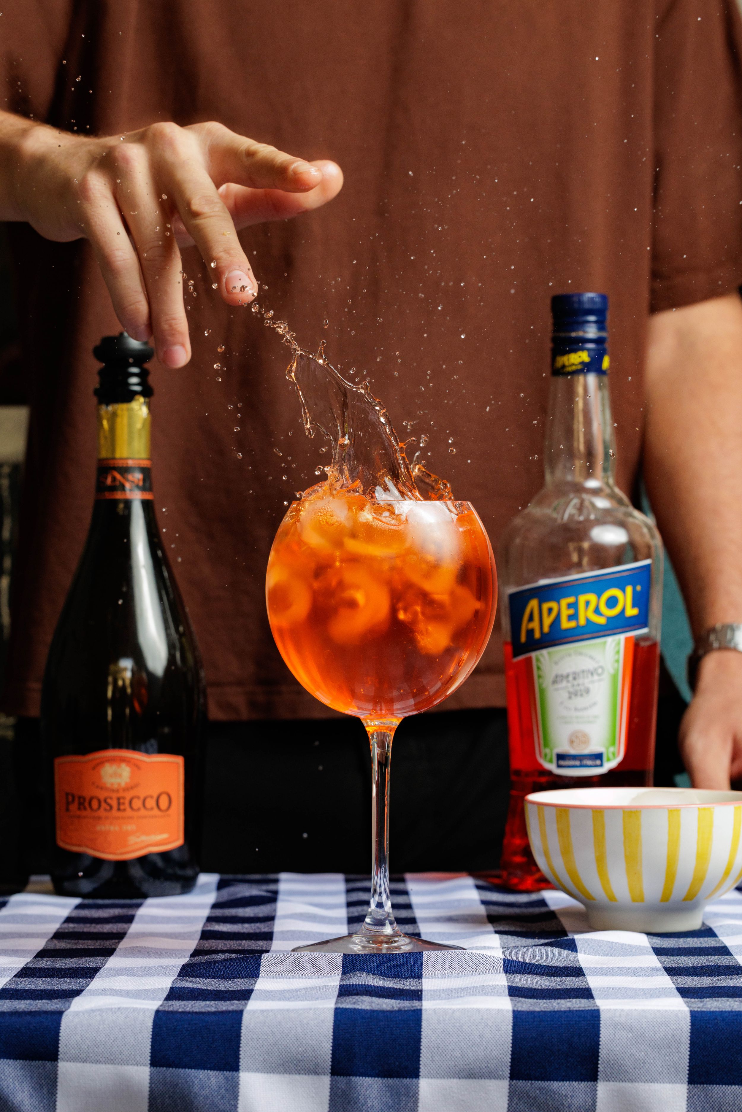 Aperol Spritz cocktails being prepared on a blue and white checkered tablecloth with Prosecco and orange garnish.