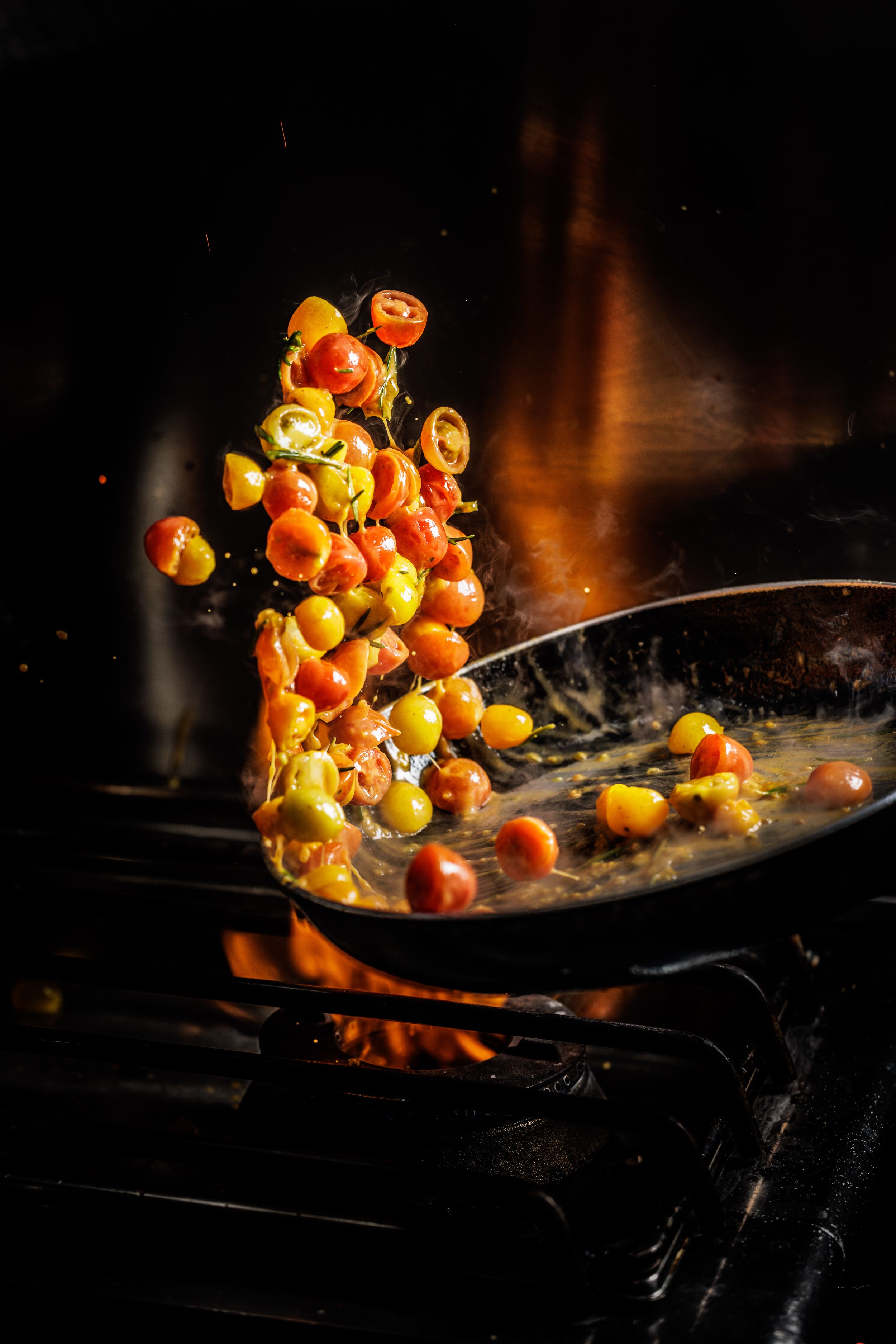 Colorful cherry tomatoes being tossed in a sizzling pan over open flames on a stovetop.