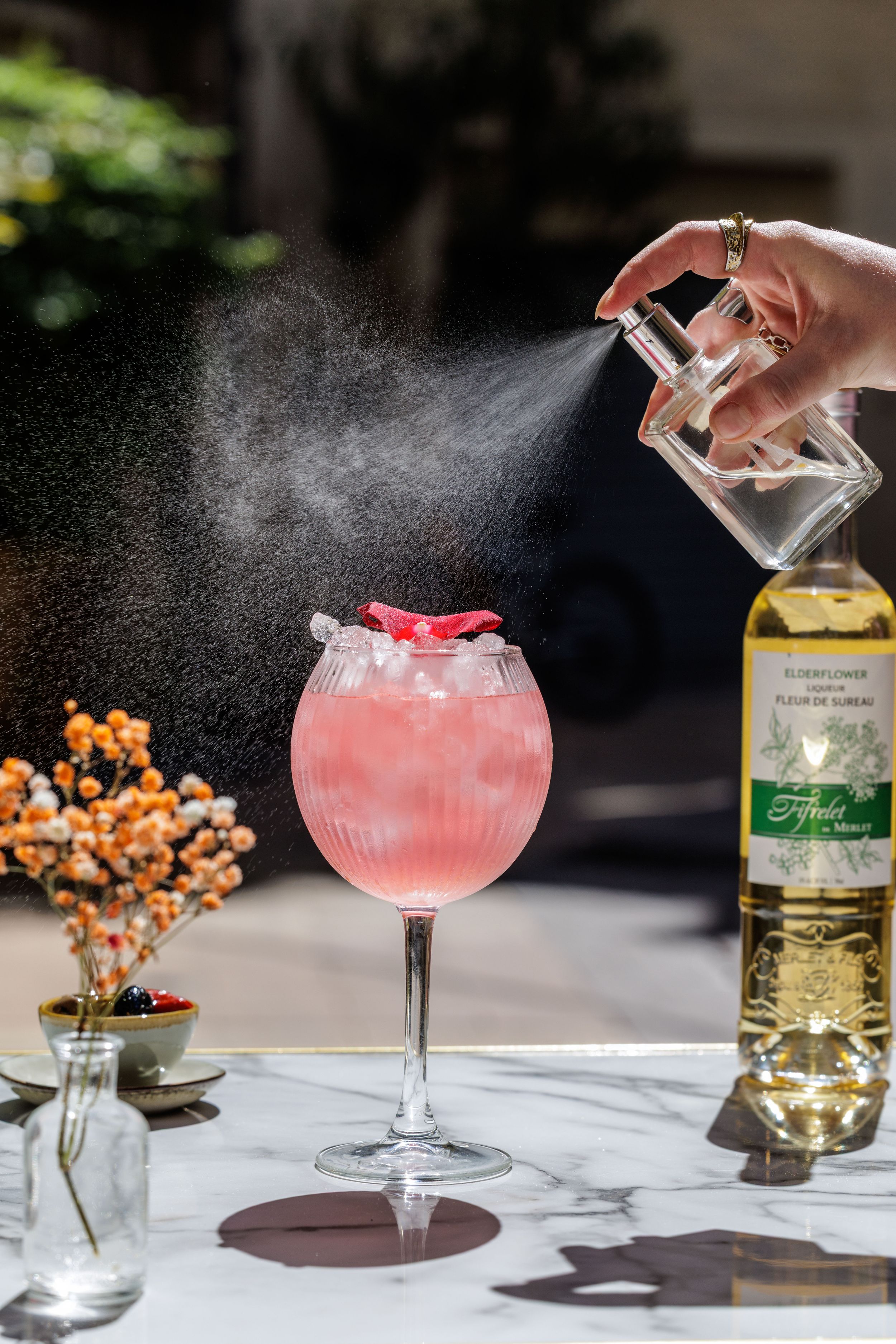 A multi-image sequence showing the preparation of a pink cocktail being garnished with sprayed mist on a marble bar top surface.