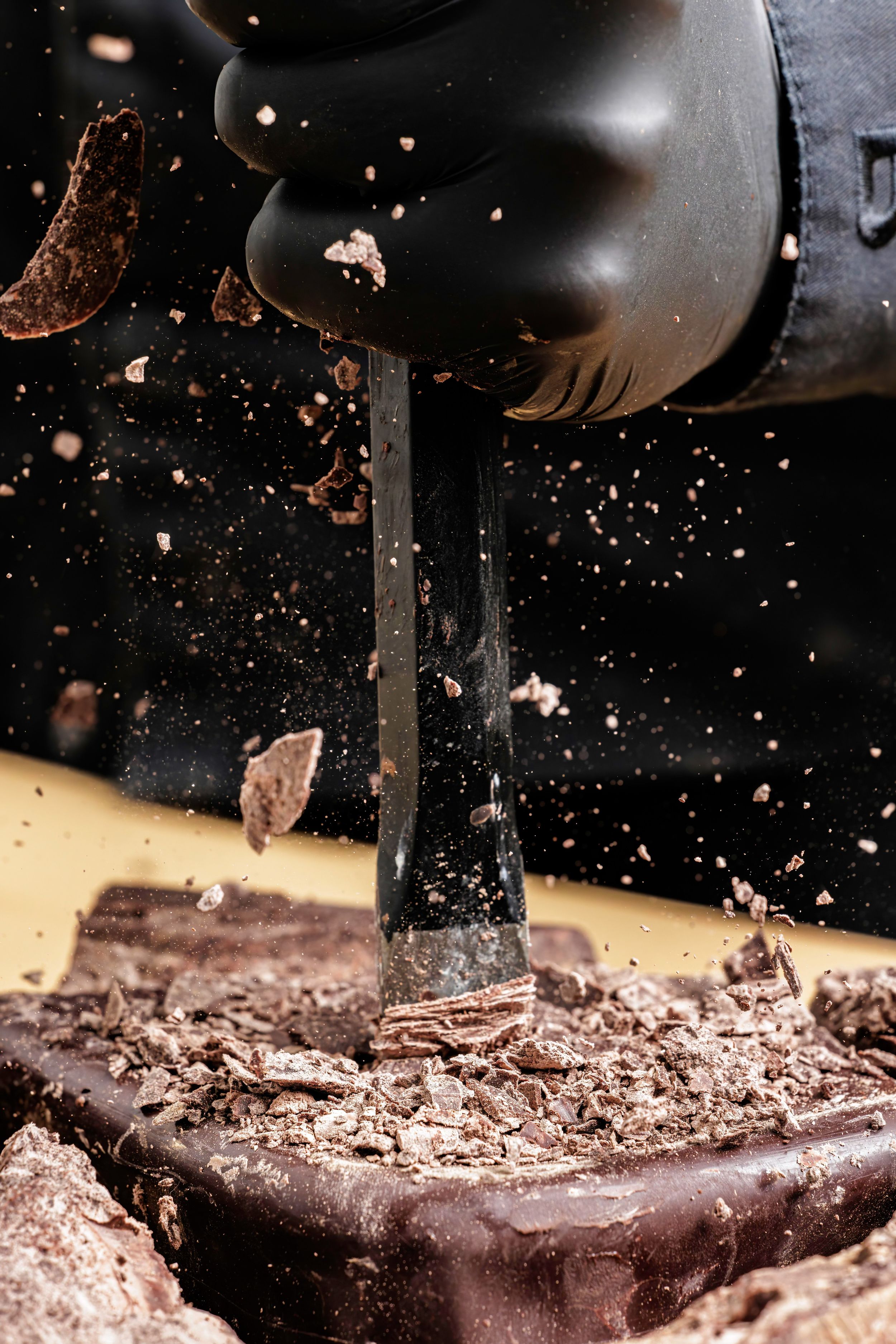 A power drill breaking through dark chocolate bar with shavings and pieces flying dramatically against black background.