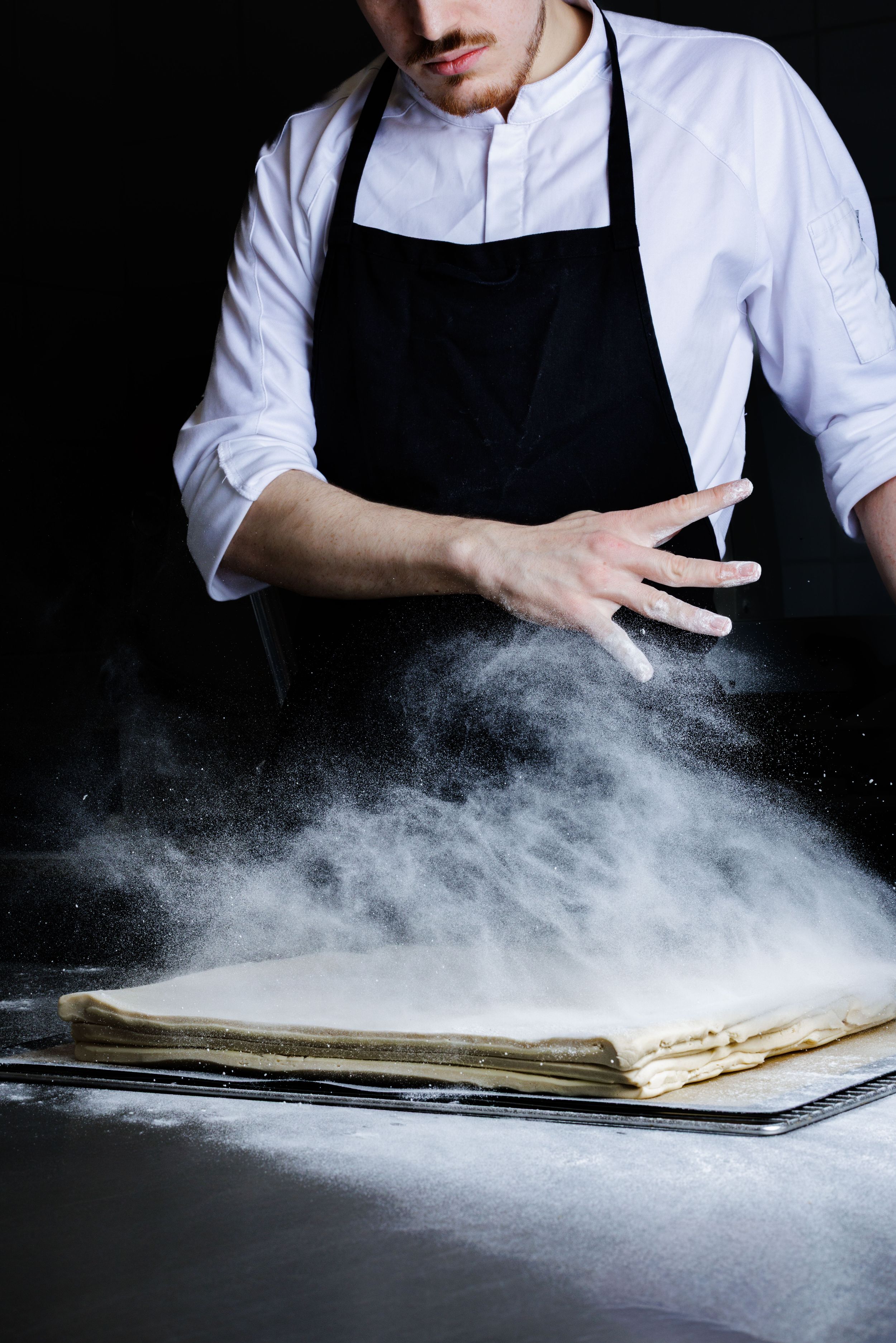 Baker in black apron creates dramatic effect by dusting flour across dark surface in sequence of action shots.