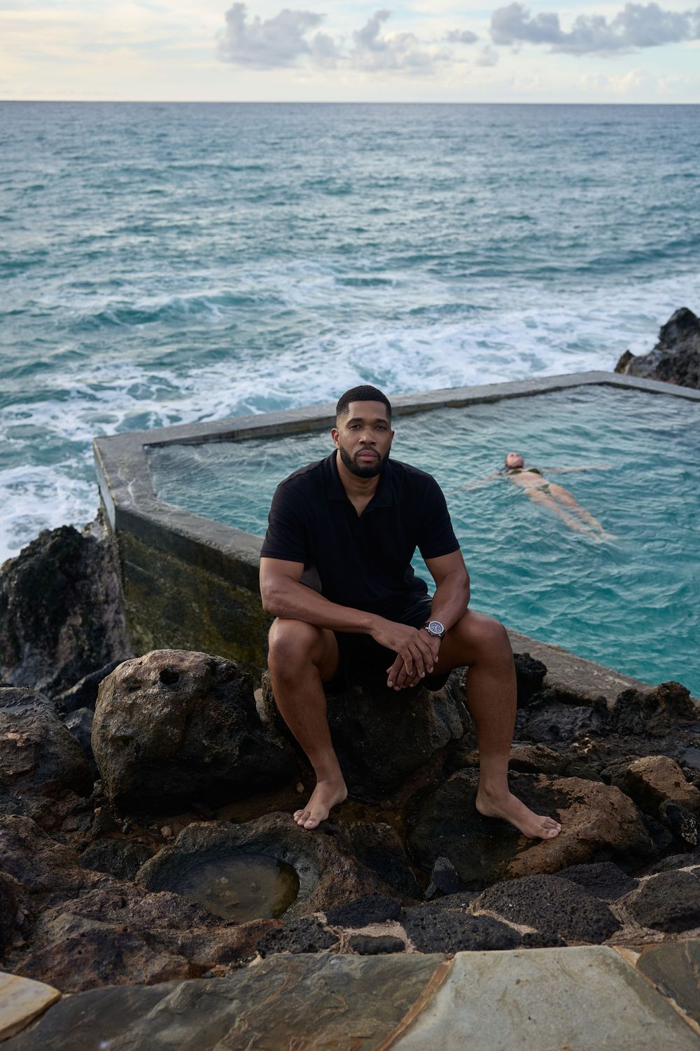 Man sitting by ocean pool at sunset, leaning against rocky cliff in black polo and shorts, peaceful and reflective vibe.