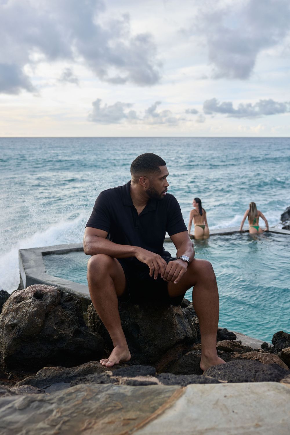 Man sitting by ocean pool at sunset, leaning against rocky cliff in black polo and shorts, peaceful and reflective vibe.